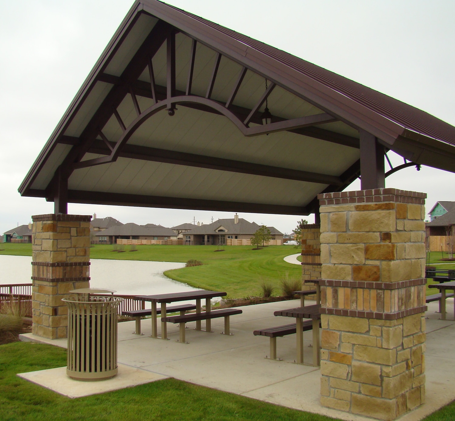 Stone and brown steel shelter with picnic benches near a lake in a neighborhood.