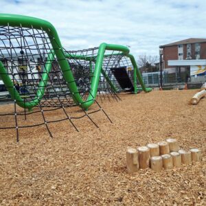 A large, abstract net climber with a thick, bright green tubular frame sits on a wood chip play surface next to small log steps.