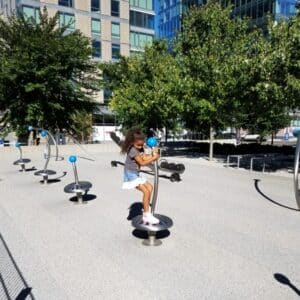 A young girl stands on a small, metallic stand-up spinner, part of a row of similar spinners in a plaza.