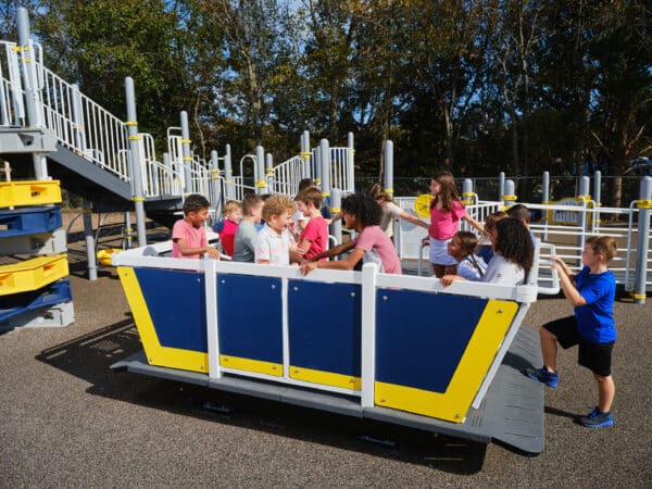 A large group of children stand and sit together inside a blue and yellow, boat-like playground glider on a sunny day.