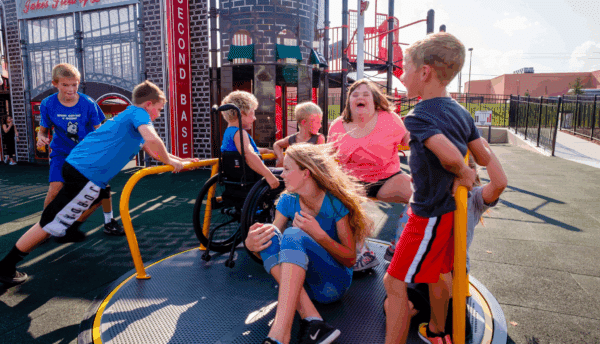 A group of smiling children gathered on an accessible carousel at a playground.