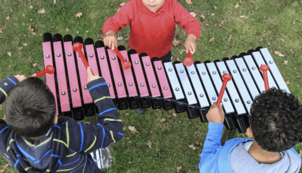 Three children seen from above play a large outdoor xylophone with red and silver keys on a grassy lawn.