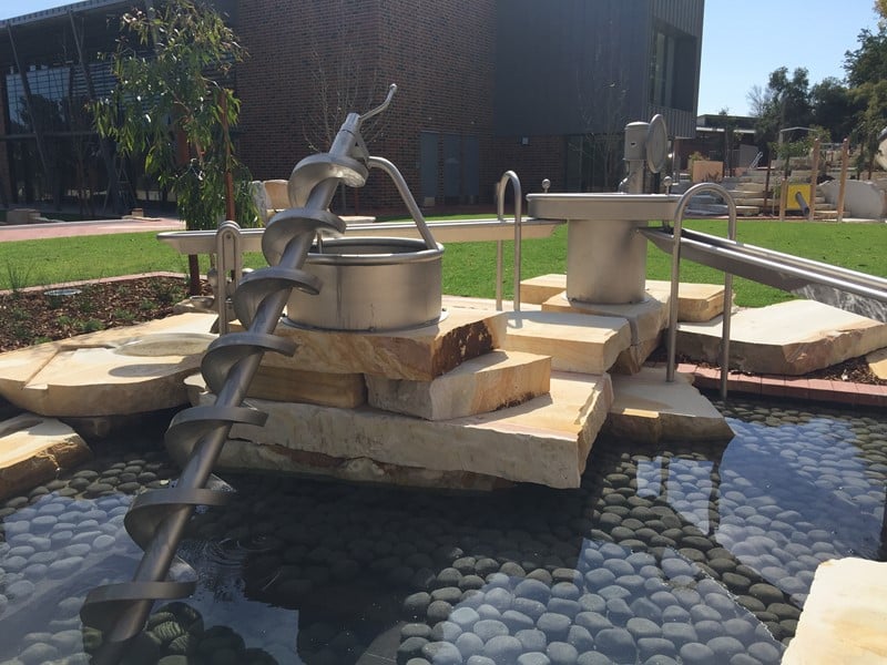 Stainless steel Archimedes screw water play feature mounted on sandstone blocks, lifting water from a pebble basin.