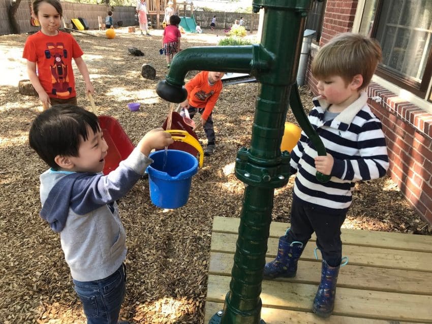 Two young children using a green water pump on a playground; one pumps the handle while the other fills a blue bucket.