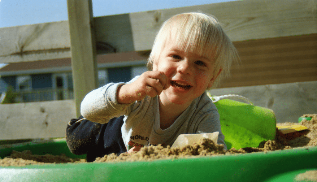 Smiling toddler lying in a green sandbox, holding a handful of sand during outdoor sensory play.