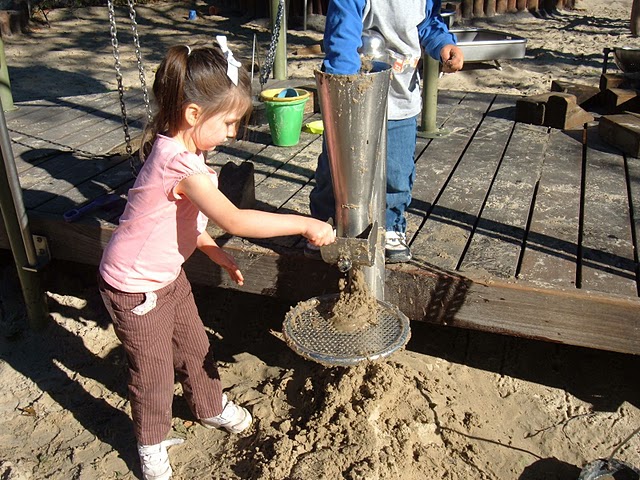 Young girl playing with a stainless steel sand silo and sieve tray feature on a wooden playground deck.