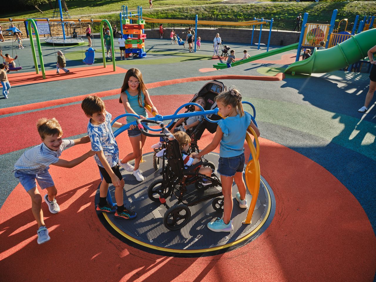 Children spin a wheelchair-accessible playground carousel with a child in a mobility chair at an inclusive playground.