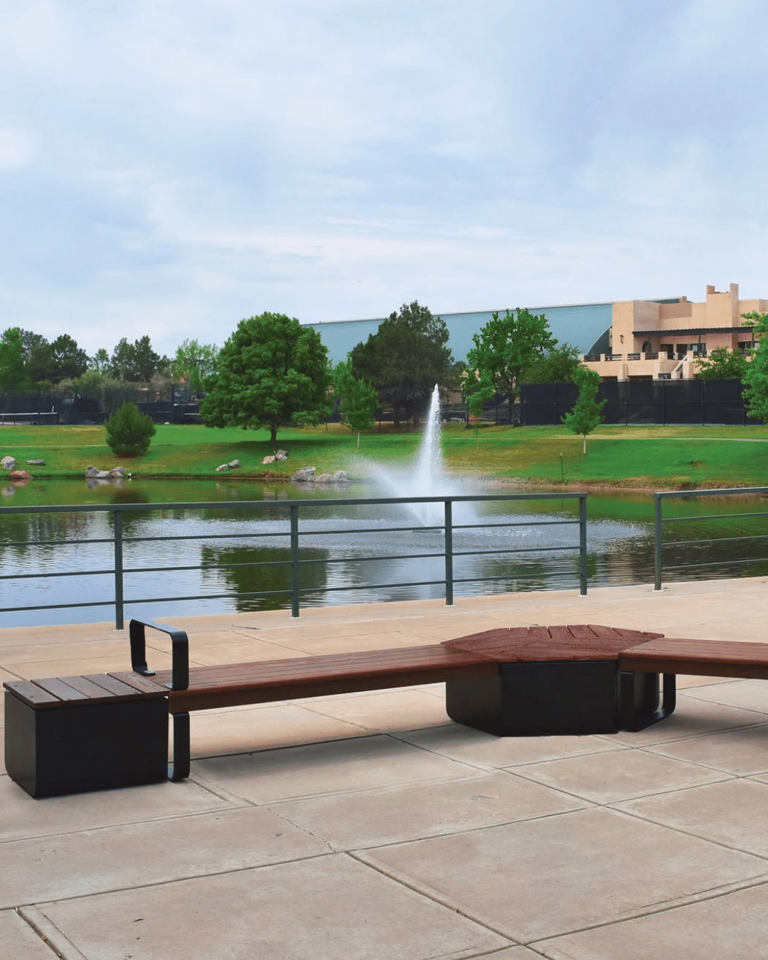 A long modular outdoor bench with dark metal base pedestals and warm wood-slat seating, positioned on a concrete plaza overlooking a pond with an active water fountain and campus buildings in the background.