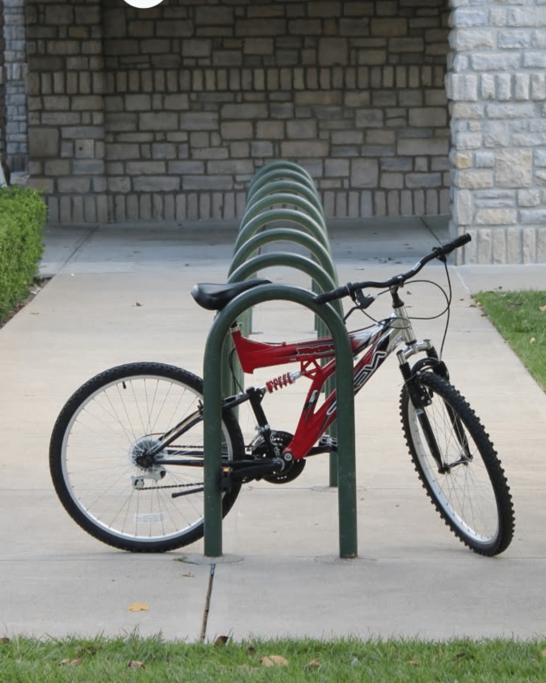 A red mountain bike locked to a green tubular loop bike rack installed along a concrete walkway in front of a stone building.