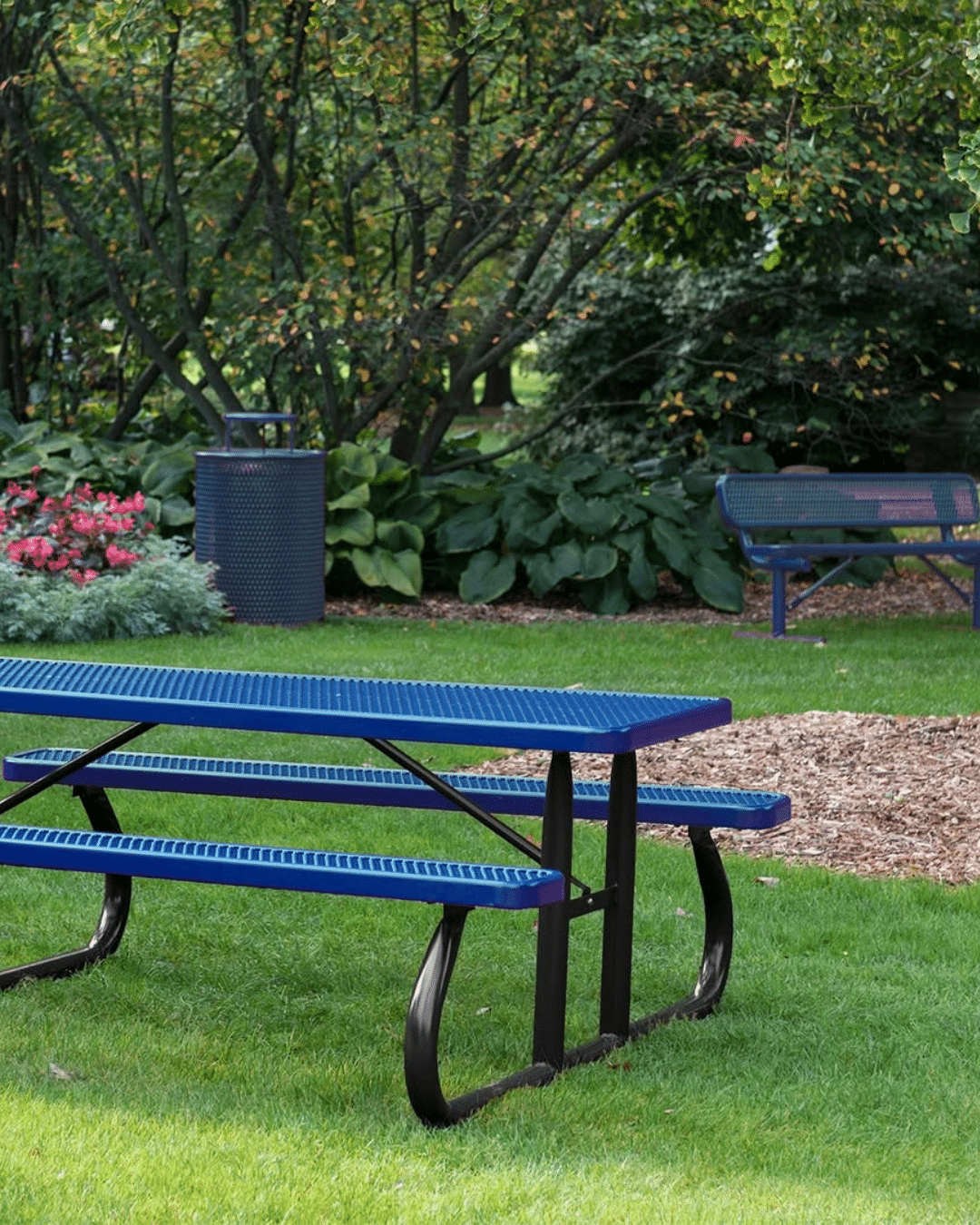 A blue metal mesh picnic table set on a green lawn, with a matching blue trash receptacle and a backless park bench visible in the background against lush foliage.
